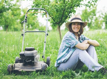 lady in a hat sitting on her lawn smiling next to her lawnmower